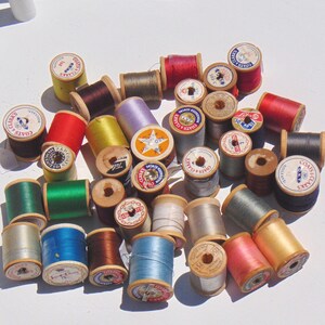 May include: A collection of assorted sewing thread spools with wooden cores, featuring various solid colors including red, blue, green, yellow, and metallic tones. Each spool has a circular paper label on the end with brand text like Coats & Clark.