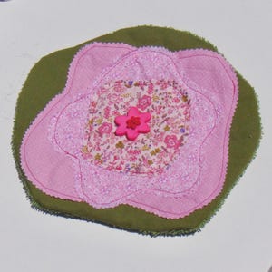 May include: A fabric coaster featuring a layered flower design with a solid olive green base, a light pink polka dot petal layer, and a central floral print fabric patch. A bright pink flower-shaped button is stitched to the center.