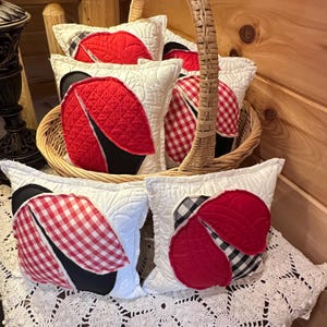 May include: A collection of decorative pillows shaped like ladybugs. The pillows have white quilted backgrounds with red, black, and gingham accents. Some are in a wicker basket, others are on a white lace surface.