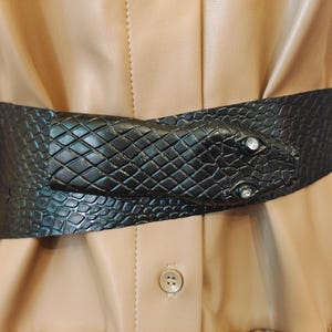 Op de afbeelding: Een zwarte riem met een slangvormige gesp. De riem is gemaakt van een textuurrijk materiaal en heeft twee heldere, ronde versieringen voor ogen. De riem is om een ​​beige jurk gewikkeld.