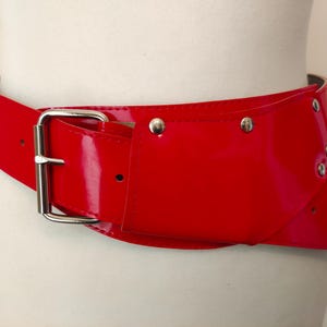 Op de afbeelding: Een brede, glanzende rode riem met een zilverkleurige gesp en klinknagels. De riem is gemaakt van glanzend materiaal en is ontworpen om de taille te accentueren. De gesp is vierkant en de riem heeft een klassiek ontwerp.