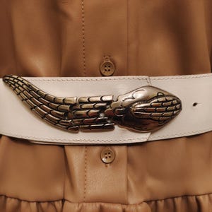 May include: A white leather belt with a silver snake-head buckle. The belt is worn over a tan leather dress. The snake buckle is detailed with scales and a curved design. The dress has buttons and a ruffled hem.