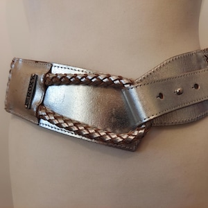 Puede incluir: Un cinturón de cuero plateado con un diseño trenzado y una hebilla plateada que dice "ROXY".