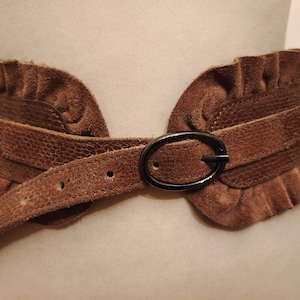 Puede incluir: Un cinturón de ante marrón con un diseño ancho y con volantes. El cinturón presenta un panel central texturizado y una hebilla ovalada oscura. El color del cinturón es un marrón cálido y terroso, y parece ser de cuero.