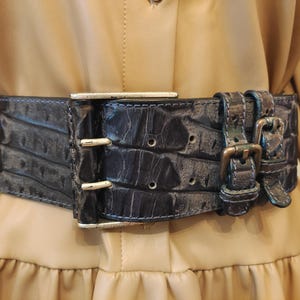 Op de afbeelding: Een brede, donkerblauwe riem met een gestructureerd, in reliëf gemaakt krokodillenpatroon. De riem heeft een grote, rechthoekige zilverkleurige metalen gesp met twee pinnen en twee kleinere, bijpassende gespen aan de zijkant voor verstelling.