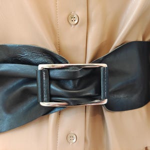 Op de afbeelding: Een brede zwarte riem met een geplooide, gerimpelde textuur en een rechthoekige zilverkleurige metalen gesp. De riem wordt gedragen over een lichtbeige overhemd met zichtbare knopen, wat een contrast vormt tussen het donkere accessoire en het lichte kledingstuk.