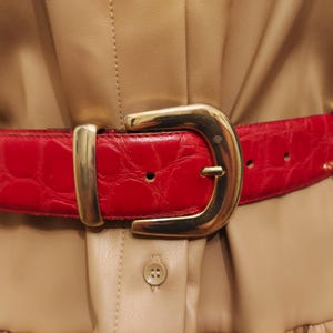 Può includere: Primo piano di una cintura rossa con fibbia dorata. La cintura presenta un motivo strutturato, simile alla pelle di coccodrillo. La fibbia dorata è grande e rettangolare. La cintura è indossata su un vestito beige.
