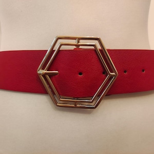 Peut inclure: Large ceinture rouge avec une boucle géométrique dorée. La ceinture est faite d'un matériau lisse et comporte trois trous de réglage. La boucle est une grande conception ouverte en forme hexagonale.