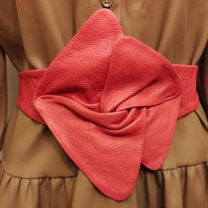May include: A vibrant red leather belt with a distinctive knot detail, worn over a brown dress. The belt provides a striking contrast and adds visual interest. The dress features a button fastening and a ruffled skirt.
