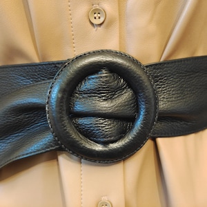 Può includere: Una cintura in pelle nera con una grande fibbia rotonda. La cintura è avvolta attorno a un capo beige con dettagli di bottoni. La fibbia è il punto focale, con una superficie strutturata e un design circolare.
