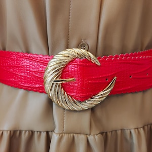 Può includere: Una cintura rosso brillante con una trama testurizzata simile alla pelle di rettile e bordi smerlati. La cintura presenta una grande fibbia circolare in metallo dorato con un design ritorto e scanalato che fissa il cinturino con un perno.