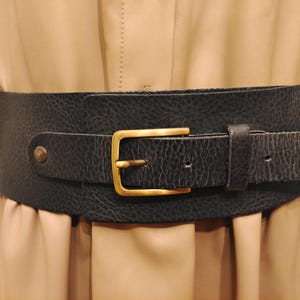 May include: A wide black leather belt with a textured finish and a gold-coloured rectangular buckle. The belt is worn over a tan dress, creating a cinched waist. The belt has a single belt loop and a brass-coloured stud.
