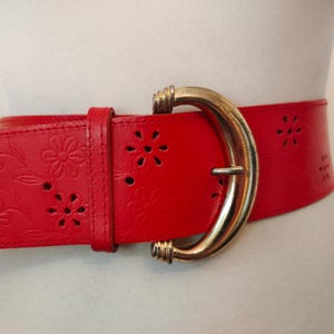 Peut inclure: Large ceinture en cuir rouge avec une boucle dorée. La ceinture présente un motif floral ajouré et est attachée autour d'un mannequin. La boucle est de forme incurvée.