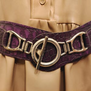 May include: A wide, purple belt with a textured, crocodile-like pattern. The belt features a large, silver-colored buckle with a circular design and a bar closure. The belt is worn over a tan-colored dress.
