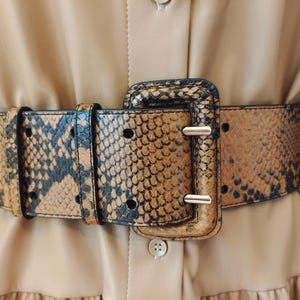 Op de afbeelding: Een brede riem met een bruin en zwart slangenleerpatroon en een bijpassende rechthoekige gesp. De riem heeft twee rijen ronde gaten over de lengte en wordt gedragen over een effen beige kledingstuk met een zichtbare knoop en naad.