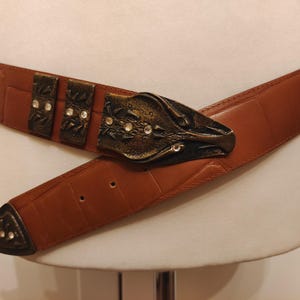 May include: A brown leather belt with a decorative metal buckle. The buckle has an ornate design with clear crystal accents. The belt is displayed on a mannequin and has a textured surface.