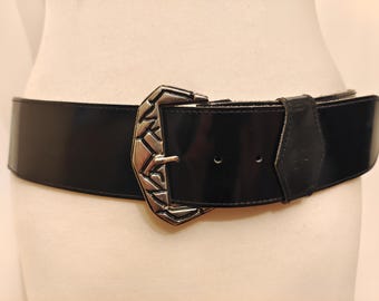 May include: A wide black belt with a shiny finish and a large silver-colored buckle. The buckle has a geometric design. The belt is displayed on a mannequin.
