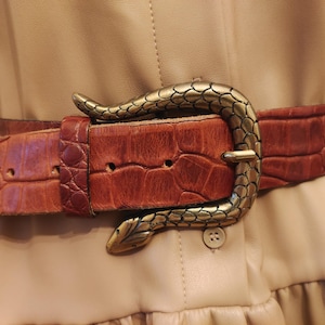 Op de afbeelding: Een bruine leren riem met een textuurpatroon dat op krokodillenleer lijkt. De riem heeft een decoratieve, antiek goudkleurige slangenriemgesp. De riem wordt over een beige jurk gedragen.