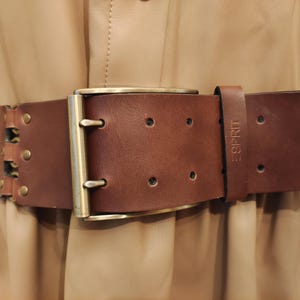 Può includere: Un'ampia cintura in pelle marrone con una grande fibbia color oro. La cintura ha un design rettangolare con rivetti e la scritta "ESPRIT" in rilievo sul cinturino. La cintura è indossata sopra un vestito beige.