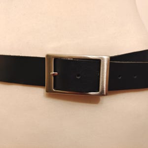 May include: A black leather belt with a silver-colored square buckle. The belt is worn around a mannequin's waist. The belt has a simple, classic design.