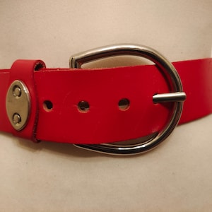 Può includere: Una cintura in pelle rosso vivo con una grande fibbia color argento. La cintura ha un design classico con una fibbia arrotondata e una serie di fori per la regolazione. La cintura è mostrata su un manichino.