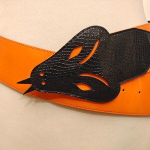 Könnte beinhalten: Ein breiter orangefarbener Ledergürtel mit einer schwarzen Lederschnalle in Form eines Vogels. Die Schnalle hat ein ausgeschnittenes Design und eine strukturierte Oberfläche.