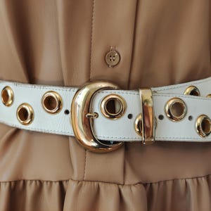 Op de afbeelding: Een witte leren riem met grote, goudkleurige metalen oogjes die gelijkmatig over de band zijn verdeeld. De riem wordt vastgemaakt met een grote, halvemaanvormige goudkleurige metalen gesp en een bijpassende metalen lus, gedragen over een beige jurk.