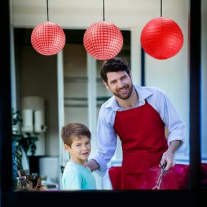 May include: A man and a boy are standing in front of a grill. The man is wearing a red apron and the boy is wearing a blue shirt. There are red and white checkered paper lanterns hanging in the background.