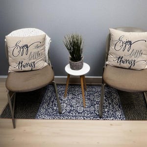 May include: Two chairs with tan cushions and metal frames flank a small white table with a woven basket and faux greenery. Each chair has a decorative pillow with the text "Enjoy the little things". A patterned rug sits beneath.