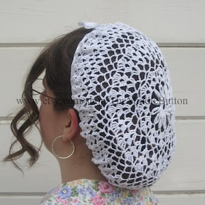 May include: White crocheted hairnet with a delicate floral pattern. The hairnet is worn by a person with brown hair and is secured with a white ribbon.