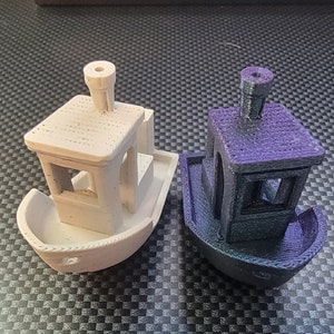 Könnte beinhalten: Zwei kleine, 3D-gedruckte Modellboote. Eines ist weiß und das andere ist dunkelviolett. Beide haben eine kleine Kabine mit einem Schornstein auf dem Dach.