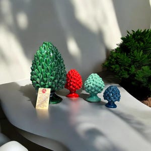 Può includere: Quattro sculture di pigne in ceramica di diverse dimensioni. La più grande è verde, la successiva è rossa, poi verde e la più piccola è blu.
