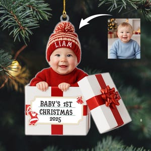 May include: A personalized Christmas ornament featuring a baby wearing a red hat with the name "Liam". The ornament includes a photo of the baby and a gift box with the text "Baby's 1st Christmas 2025". The gift box has a red bow.