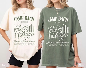 May include: Two t-shirts with a camping theme. The shirts are in ivory and moss green. The text on the shirts reads "Camp Bach Est. 2023" and "Jessica's Bachelorette Lake Tahoe, California".