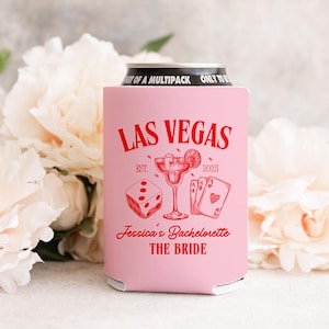 May include: A pink can cooler with red text and graphics. The text reads "LAS VEGAS" above a dice, cocktail, and playing cards. Below, it says "Jessica's Bachelorette THE BRIDE" and "EST. 2025".