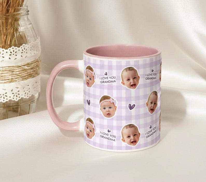 May include: A ceramic mug with a pink handle and interior. The mug features a repeating pattern of baby faces, purple hearts, and the text "I LOVE YOU GRANDMA" on a light purple gingham background. A decorative glass jar is in the background.