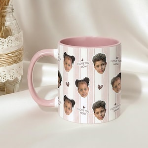 May include: White ceramic mug with a pink interior and handle. The mug features a repeating pattern of children's faces, hearts, and the text "I LOVE YOU MOM" on a striped background. A decorative jar is in the background.