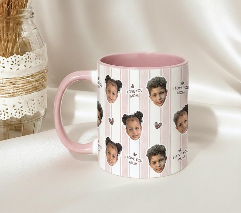May include: White ceramic mug with a pink interior and handle. The mug features a repeating pattern of children's faces, hearts, and the text "I LOVE YOU MOM" on a striped background. A decorative jar is in the background.