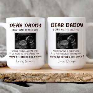 May include: Two white ceramic mugs with the text "DEAR DADDY, I CAN'T WAIT TO MEET YOU" and an ultrasound image. The mugs also have the text "YOU'RE DOING A GREAT JOB, HAPPY 1ST FATHER'S DAY, DADDY!" and "Love, Bump".