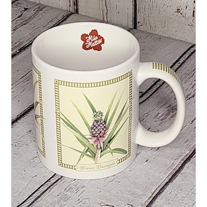 Peut inclure: Mug en céramique blanche avec une illustration d'ananas et le texte "Brunes Pineapple". La tasse a une anse avec des rayures dorées et un logo rouge qui dit "Hile Hattis".