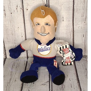 May include: Plush doll of a man wearing a white and blue racing uniform. The doll has a smiling face, brown hair, and holds a checkered flag. The text "Russ Wallace" is on the chest.