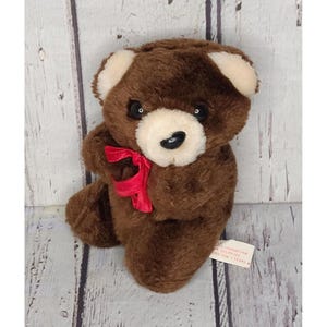 May include: A brown plush teddy bear with a red ribbon around its neck. The bear has white accents on its ears and muzzle, and black eyes. A small tag is attached to the bear.
