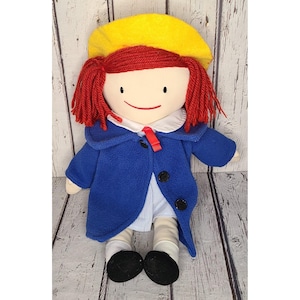 May include: A handmade rag doll with red yarn hair, a yellow hat, and a blue coat. The doll has a white collar, a red tie, and black shoes. The doll is a soft toy, perfect for children.
