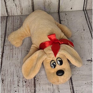 May include: A plush toy dog in a light tan color with large floppy ears and a red bow. The dog has large, round eyes and a black nose. The toy is lying on a wooden surface.