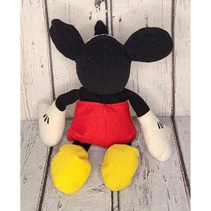 Op de afbeelding: Een pluche Mickey Mouse-speelgoed met zwarte oren, armen en een hoofd. Het speelgoed heeft een rode broek, witte handschoenen en gele schoenen. Het speelgoed staat op een lichtgekleurd houten oppervlak.