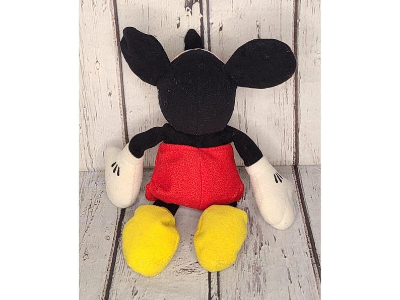 Op de afbeelding: Een pluche Mickey Mouse-speelgoed met zwarte oren, armen en een hoofd. Het speelgoed heeft een rode broek, witte handschoenen en gele schoenen. Het speelgoed staat op een lichtgekleurd houten oppervlak.