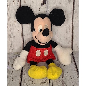 Op de afbeelding: Een pluche Mickey Mouse-speelgoed. Het iconische personage heeft grote zwarte oren, een bruin gezicht en een zwarte neus. Hij draagt een rode broek met twee witte knopen, zwarte armen, witte handschoenen en gele schoenen. Het speelgoed zit.