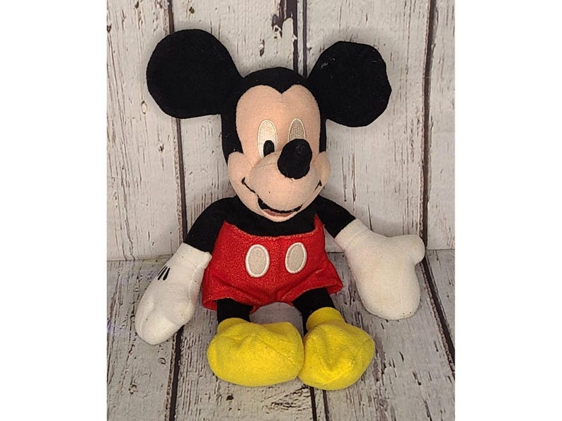 Op de afbeelding: Een pluche Mickey Mouse-speelgoed. Het iconische personage heeft grote zwarte oren, een bruin gezicht en een zwarte neus. Hij draagt een rode broek met twee witte knopen, zwarte armen, witte handschoenen en gele schoenen. Het speelgoed zit.