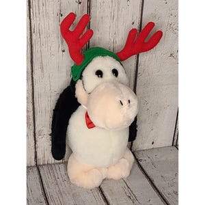 Könnte beinhalten: Ein Plüschpinguin-Spielzeug mit einem grünen Hut mit roten Geweih und einer roten Fliege. Der Pinguin hat einen weißen Körper, schwarze Flügel und ein hellrosa Gesicht und Füße. Das Spielzeug steht vor einem verwitterten, hellfarbenen Holzgrund.