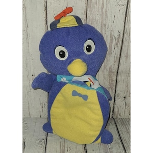 Könnte beinhalten: Ein blau-gelbes Plüschpinguin-Spielzeug mit kleinem Hut und Fliege. Der Pinguin hat große Augen und einen Schal. Das Spielzeug steht vor einem hölzernen Hintergrund.
